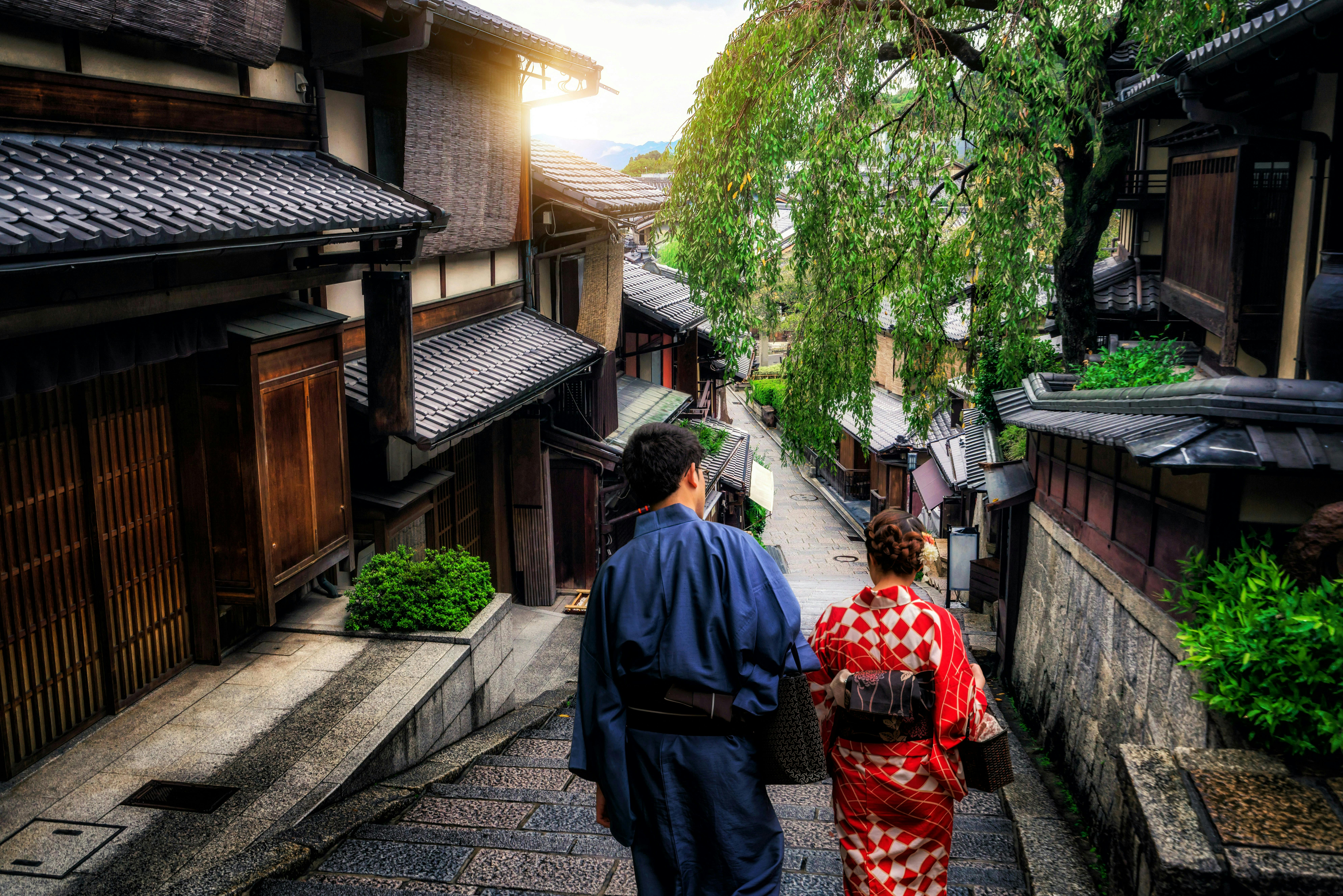 Two people in traditional Japanese clothing walk down a narrow stone street lined with wooden houses and greenery, with sunlight streaming in the background.