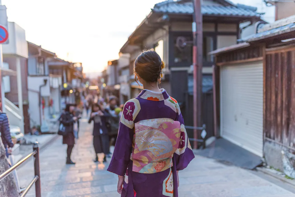 A person wearing a traditional purple kimono with floral patterns walks down a narrow, bustling street lined with wooden buildings at sunset. The setting suggests a historic or cultural area in Japan, with a blurred crowd in the background.