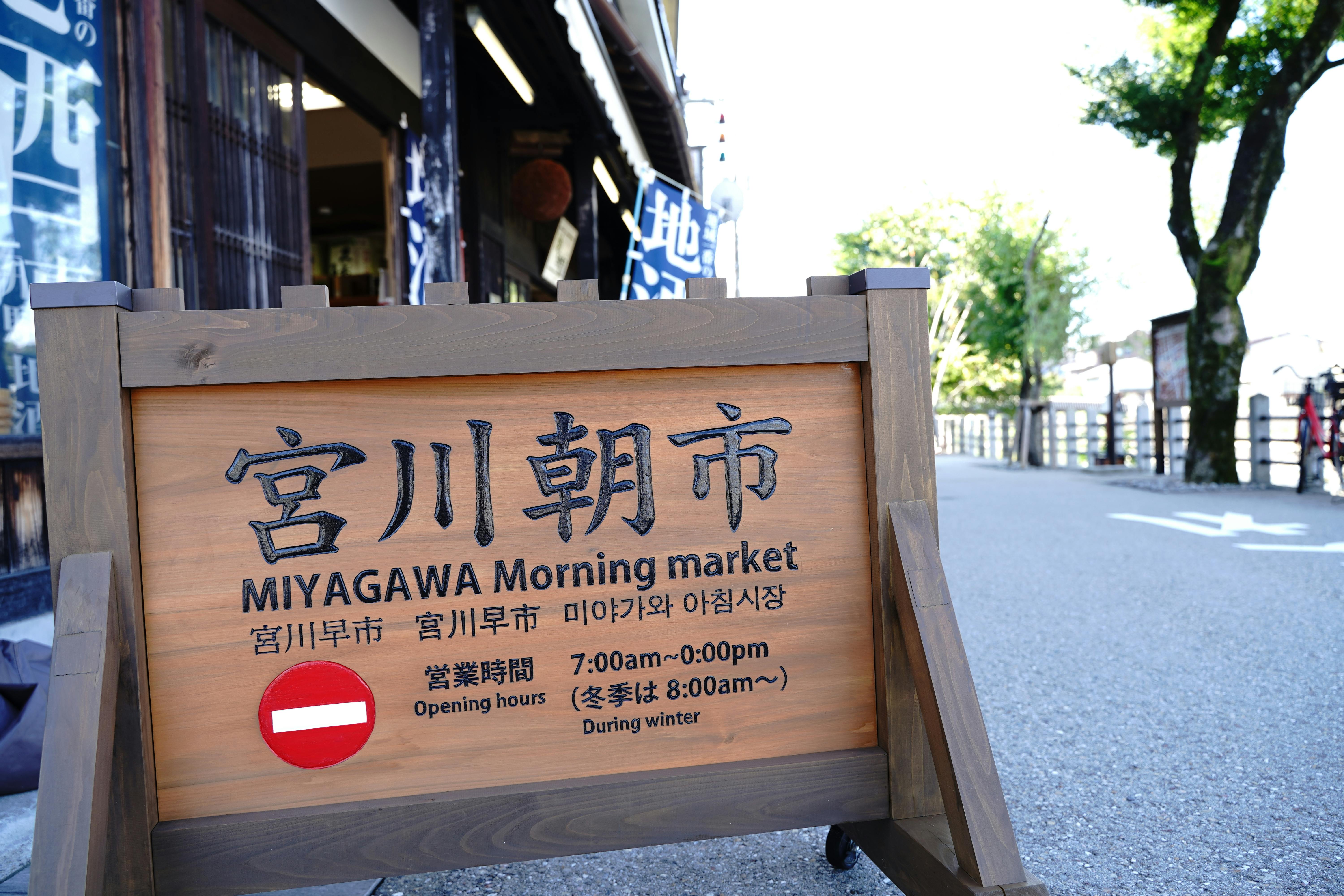 A wooden signboard reads "MIYAGAWA Morning market" with opening hours in English, Japanese, and Korean, placed on a street near trees and traditional buildings.