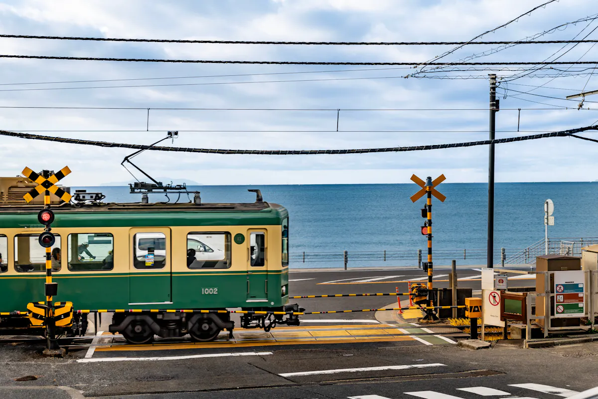 The Enoden line runs along the Shonan coast past the railroad crossing made famous by Slam Dunk. A green and cream Enoden train passing a railroad crossing with the ocean behind it on the Kamakura coast