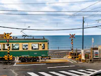 A green and cream Enoden train passing a railroad crossing with the ocean behind it on the Kamakura coast