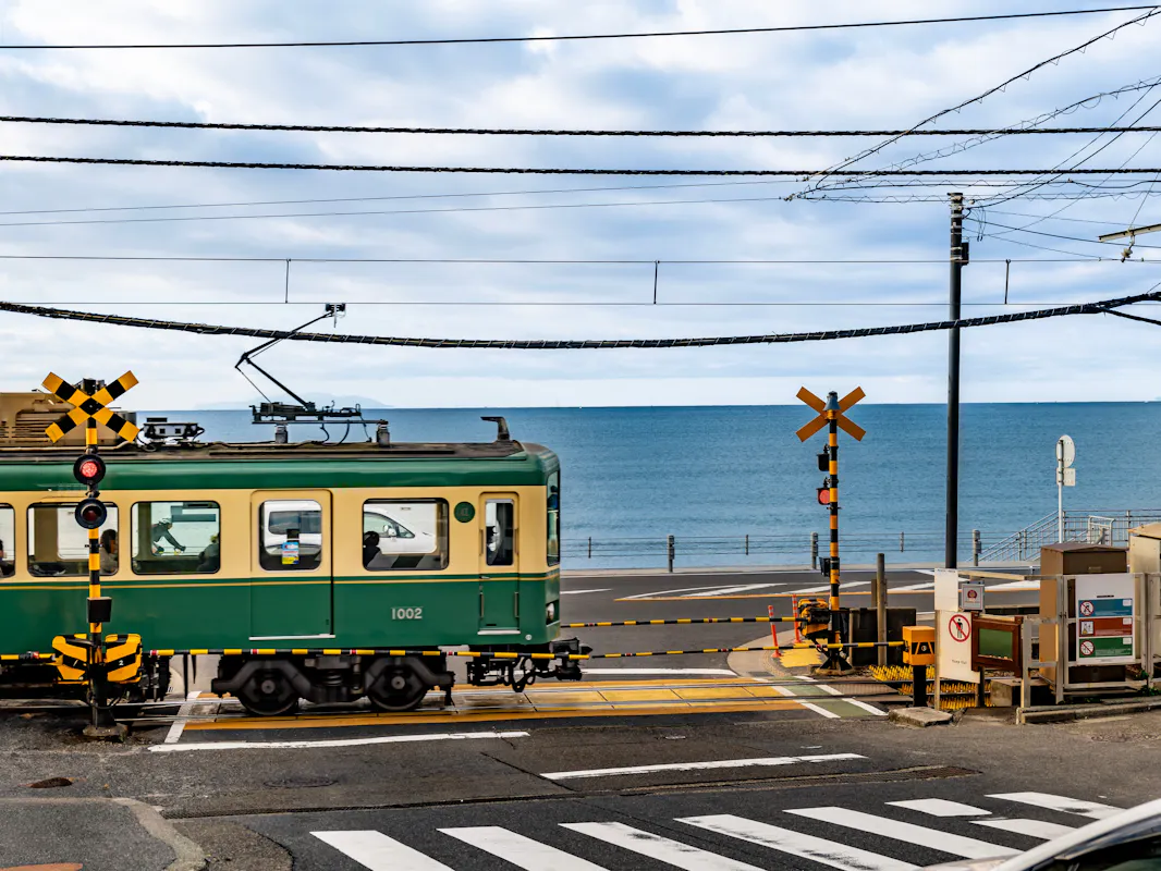 A green and cream Enoden train passing a railroad crossing with the ocean behind it on the Kamakura coast