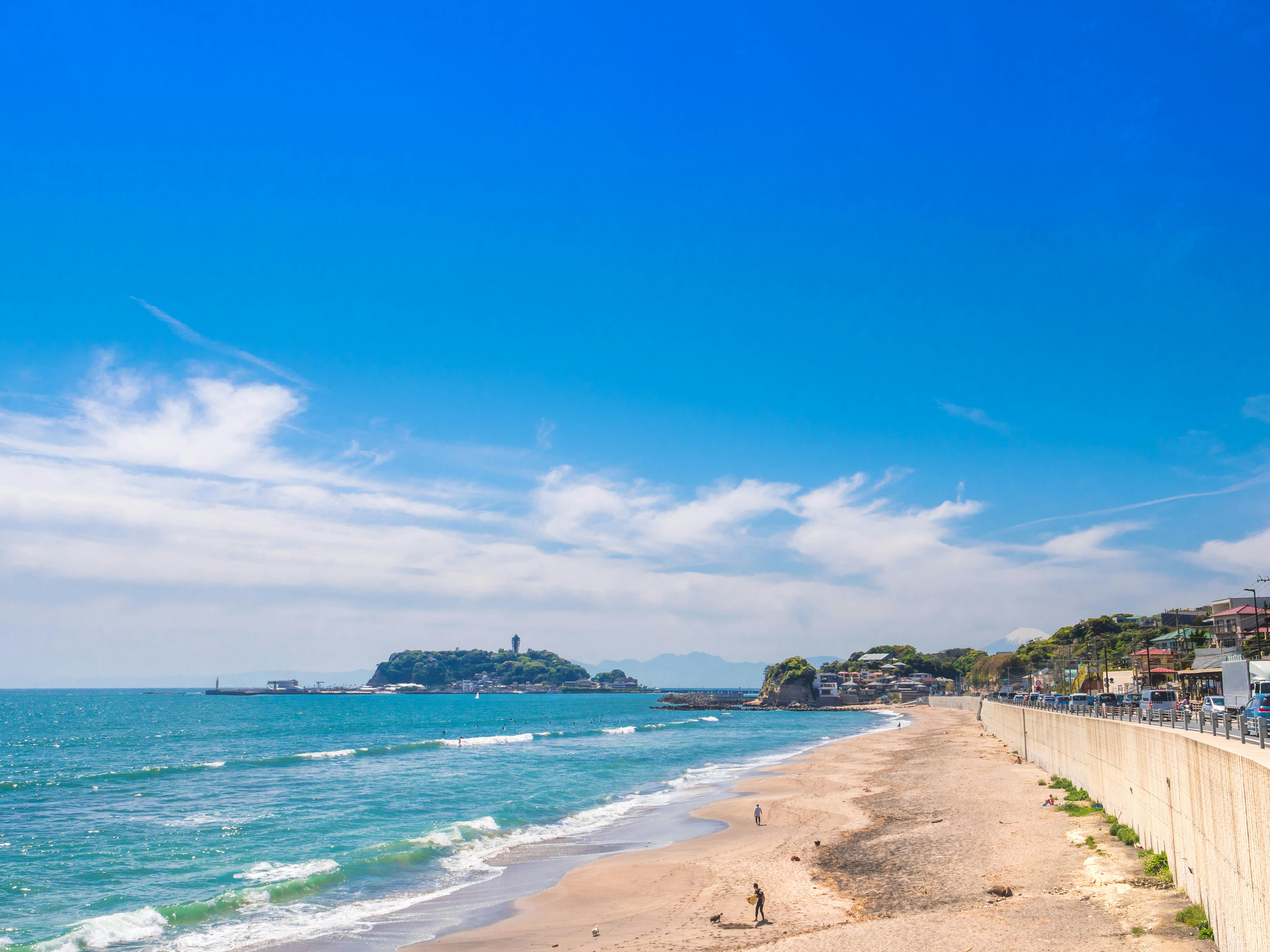 A sandy beach with gentle waves, a few people walking along the shore, and a distant island with a lighthouse under a bright blue sky dotted with wispy clouds. Houses and a wall line the right side of the beach.