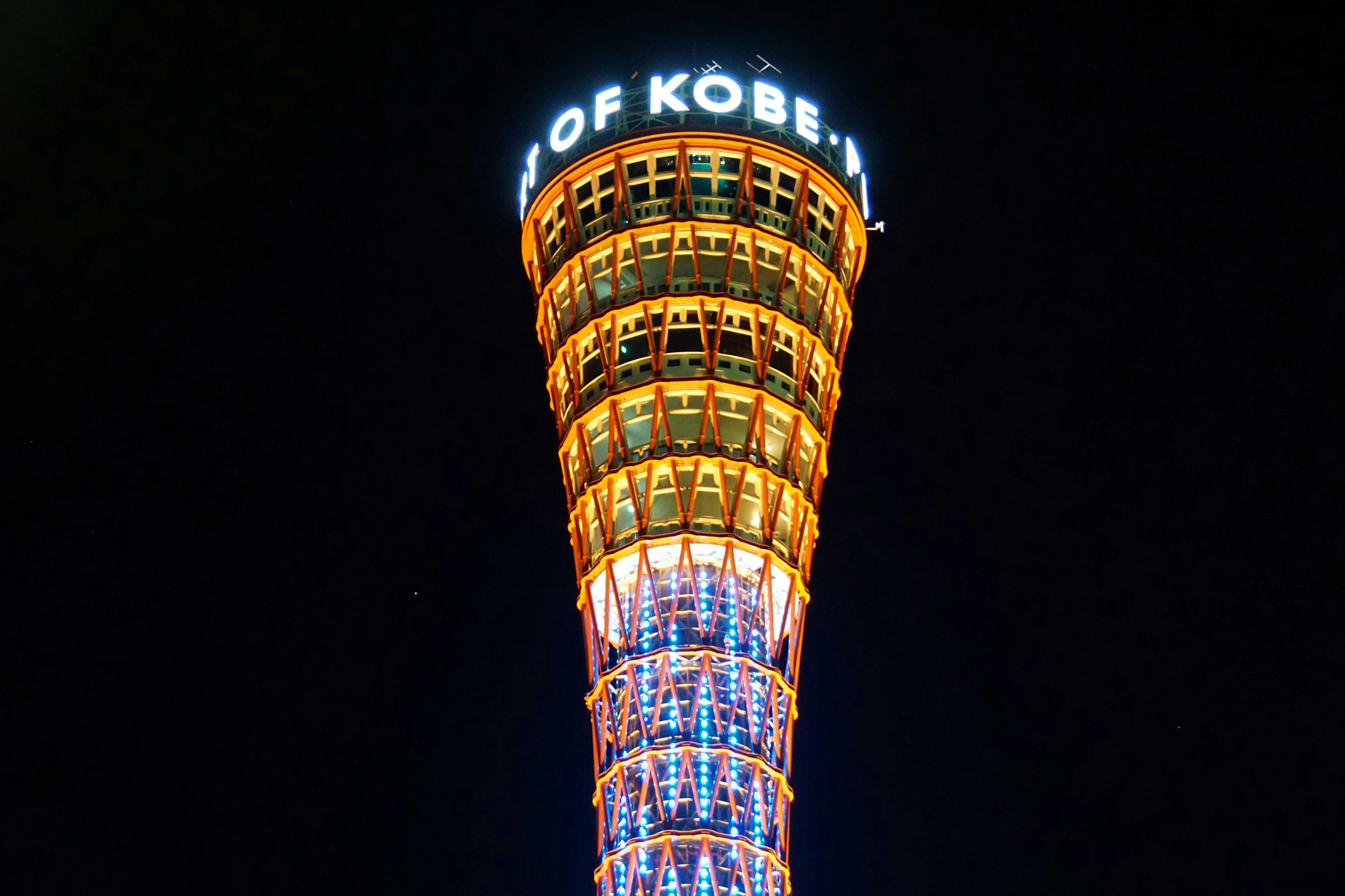 A night view of the illuminated top section of Kobe Port Tower, displaying its vibrant orange and blue lights, with part of the "KOBE" sign visible against the dark sky.