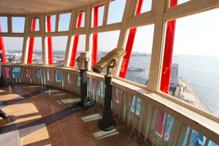 Two observation telescopes stand by large windows inside a tall tower, offering panoramic views of a coastal city and blue ocean on a bright, sunny day. Red support beams frame the windows.
