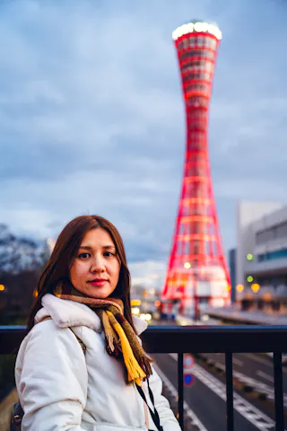 A woman in a white coat and scarf stands near a railing with Kobe Port Tower, illuminated in red lights, in the background during dusk.