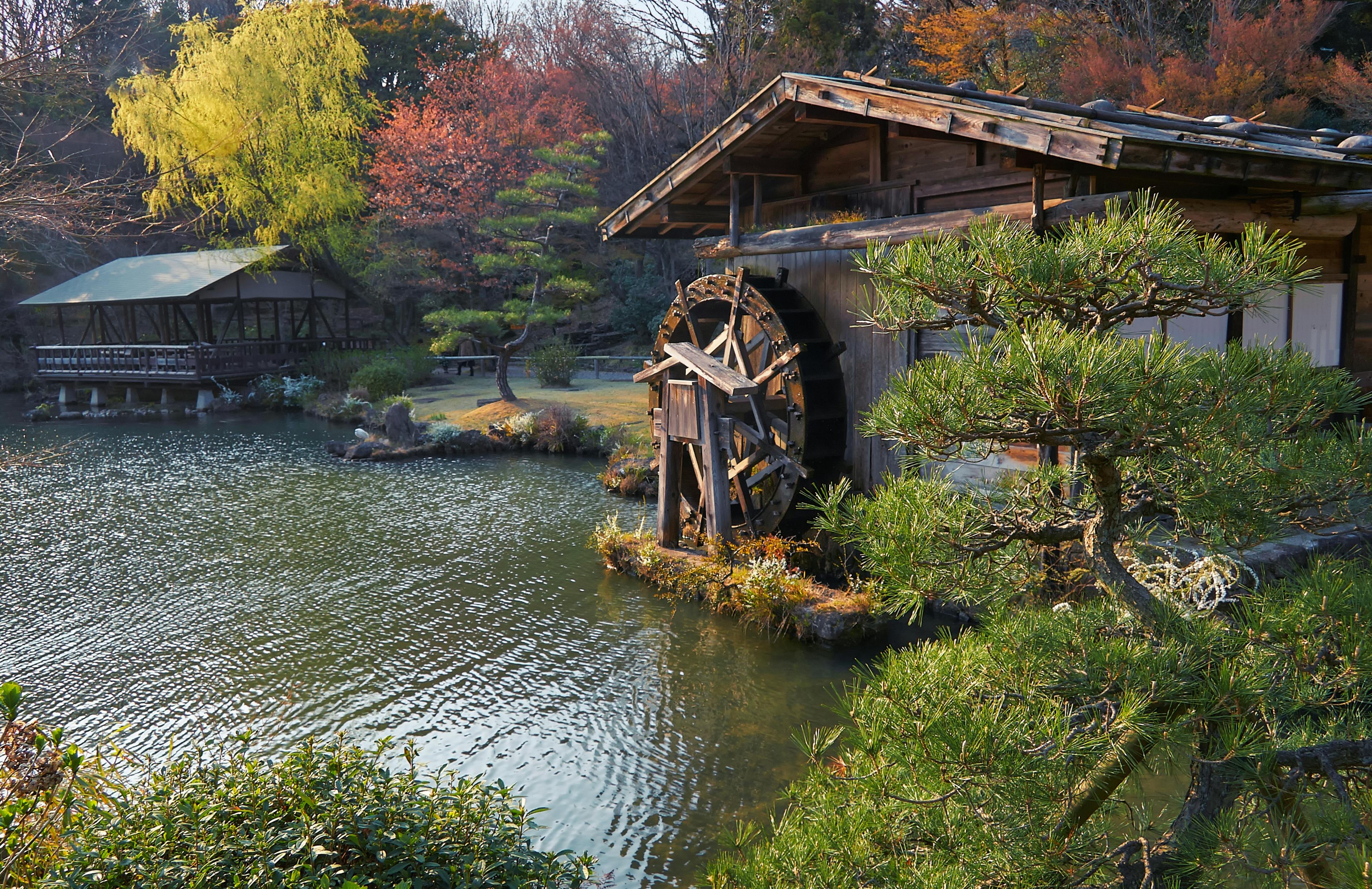 A traditional wooden watermill stands by a tranquil pond surrounded by lush greenery, pine trees, and colorful autumn foliage, with a wooden gazebo in the background.