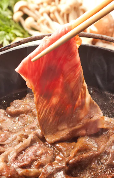 Art of Omi Beef Sukiyaki Thin slice of raw beef being cooked in a hot pot with chopsticks, surrounded by other simmering beef slices. In the background, fresh vegetables, mushrooms, and bowls with raw eggs are visible.