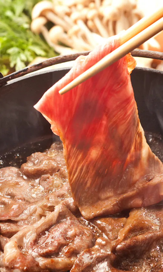 Omi Beef Sukiyaki Thin slice of raw beef being cooked in a hot pot with chopsticks, surrounded by other simmering beef slices. In the background, fresh vegetables, mushrooms, and bowls with raw eggs are visible.