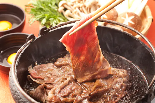 Thin slice of raw beef being cooked in a hot pot with chopsticks, surrounded by other simmering beef slices. In the background, fresh vegetables, mushrooms, and bowls with raw eggs are visible.
