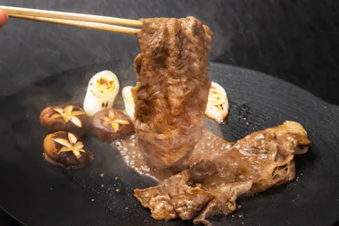 A close-up of chopsticks holding a slice of cooked beef above a plate with more beef, grilled shiitake mushrooms, and green onions, all on a dark textured background. Steam rises from the hot dish.
