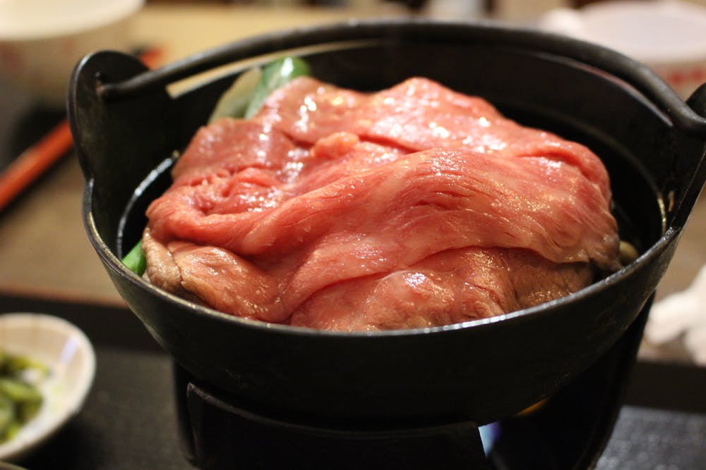 Thin slices of raw beef are arranged over vegetables in a black pot, ready to be cooked at the table. The marbled meat is fresh and pink, suggesting a Japanese hot pot or sukiyaki dish.