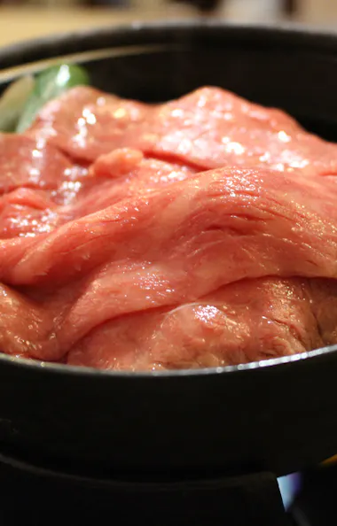 Omi Beef Sukiyaki Thin slices of raw beef are arranged over vegetables in a black pot, ready to be cooked at the table. The marbled meat is fresh and pink, suggesting a Japanese hot pot or sukiyaki dish.