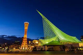 Night view of Kobe Port Tower lit in orange next to the green-lit Kobe Maritime Museum, both standing out against a deep blue sky in Kobe, Japan.