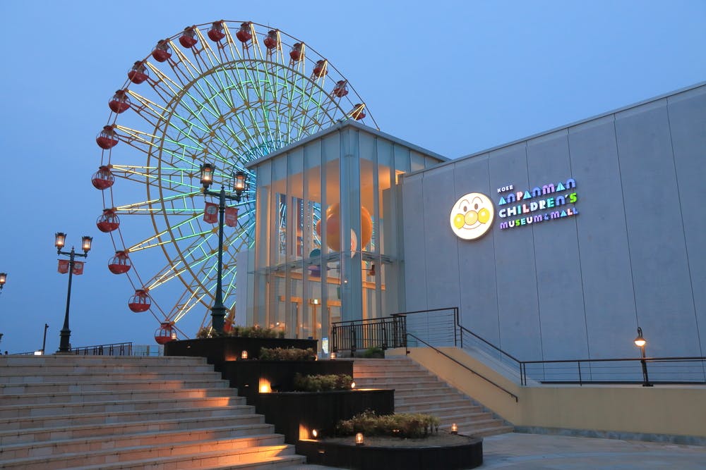 A modern building with a sign reading “Anpanman Children’s Museum & Mall” glows at dusk, with a brightly lit Ferris wheel in the background and steps with lights leading up to the entrance.