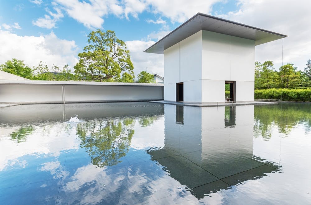 A modern white building with a large flat roof sits surrounded by a reflective pool of water, with green trees and blue sky with scattered clouds in the background.