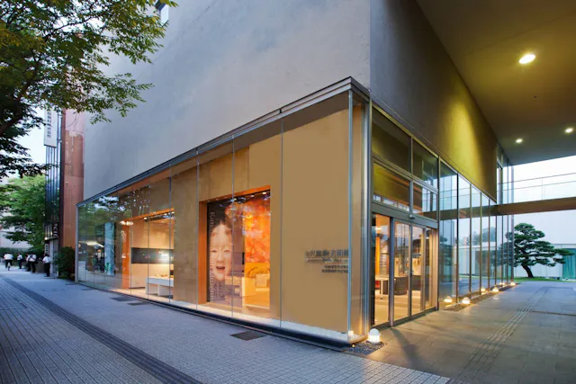 A modern glass-walled building on a tree-lined street corner, warmly lit inside with large photo displays visible through the windows; a few people walk in the background.