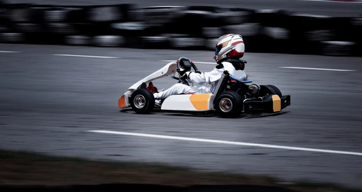 A person wearing racing gear and helmet is driving a go-kart on a race track. The image captures the speed and motion of the kart as it goes past, with blurred tires and track barriers in the background.