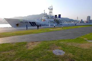 A large white coast guard ship marked "PL 05" is docked at a harbor. Several people walk and stand on the promenade near the ship, with green grass, pavement, and distant mountains in the background.