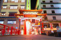 A brightly lit traditional Chinese archway with red pillars stands at the entrance to a Chinatown district, surrounded by colorful buildings at night.