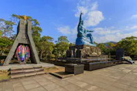 A large blue Peace Statue sits on a stone pedestal in a park, with a monument adorned with colorful paper cranes nearby. Trees and a blue sky with scattered clouds are in the background.