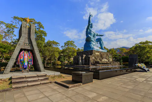 A large blue Peace Statue sits on a stone pedestal in a park, with a monument adorned with colorful paper cranes nearby. Trees and a blue sky with scattered clouds are in the background.