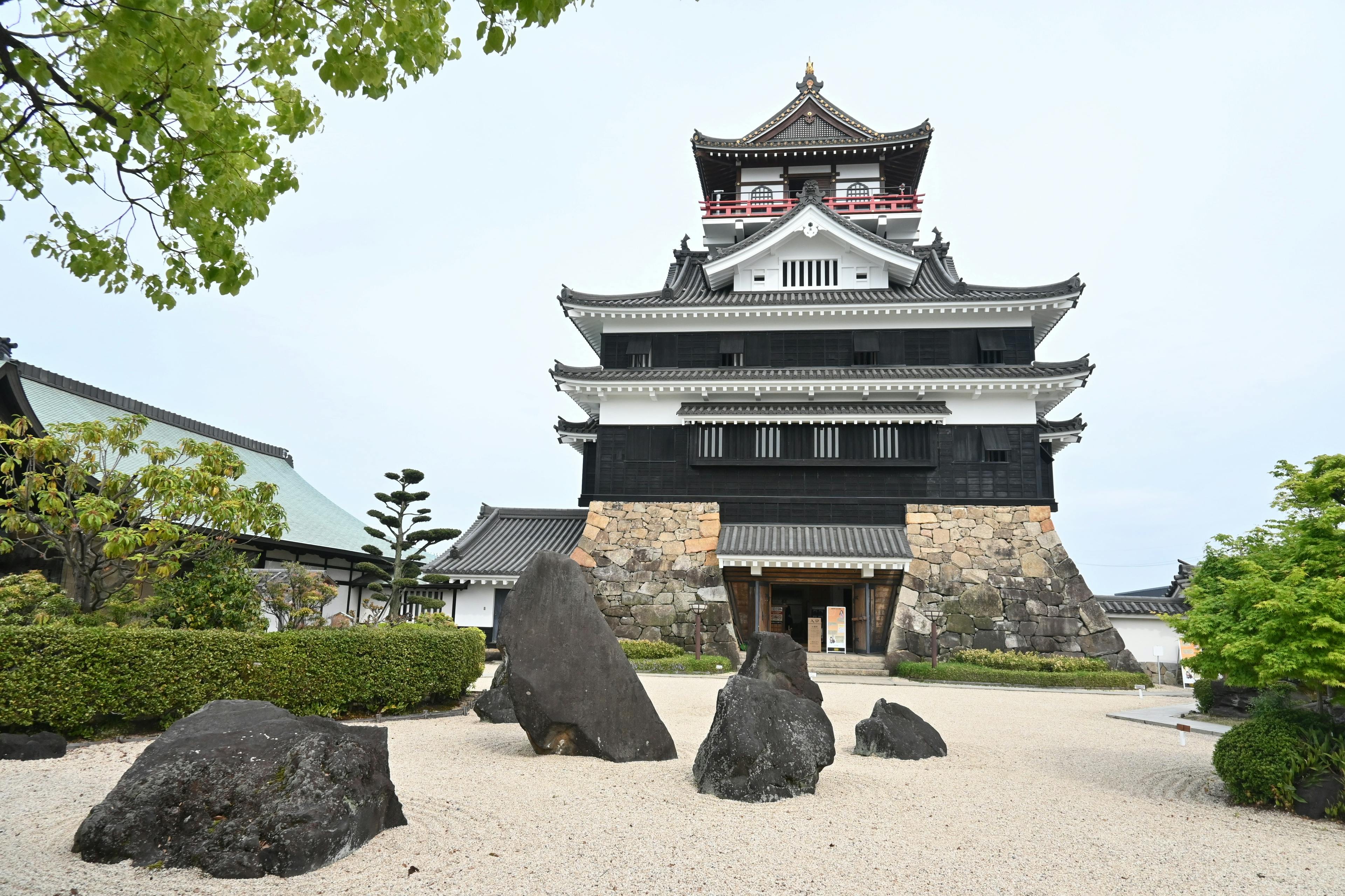 A traditional Japanese castle with white walls and black accents stands behind a rock garden with large stones and neatly trimmed shrubs, under a partly cloudy sky.