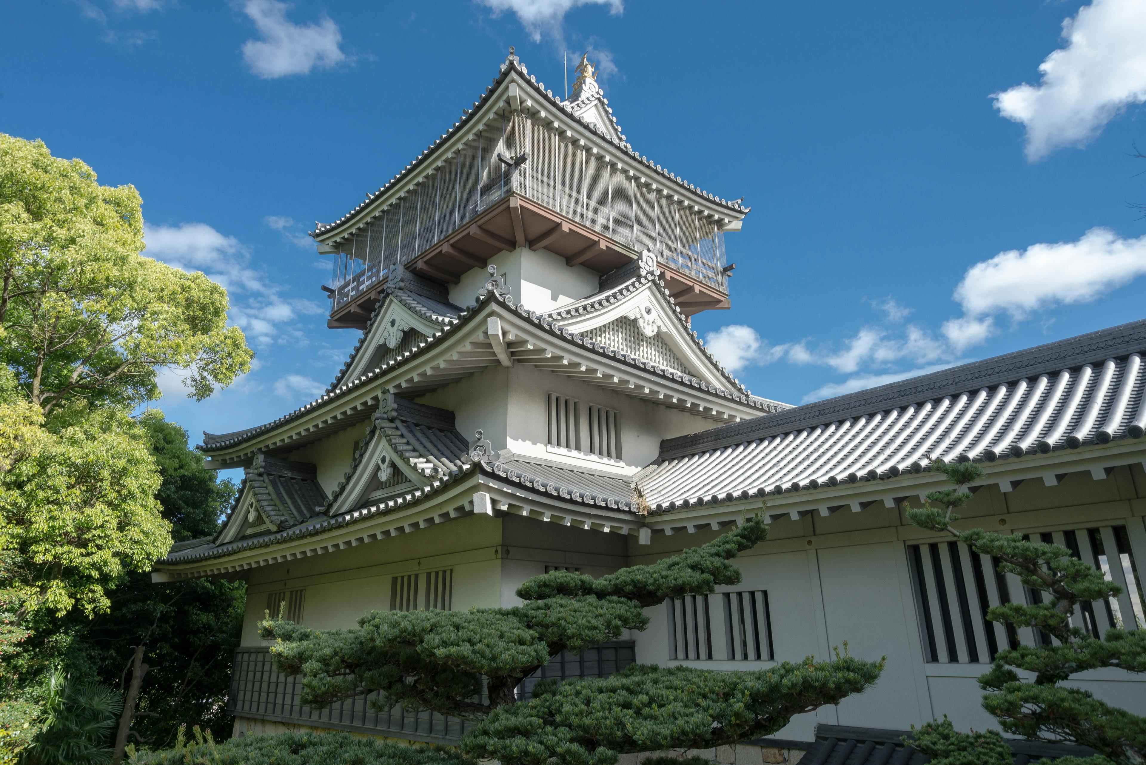 A traditional Japanese castle with white walls and tiled roofs stands under a bright blue sky with scattered clouds, surrounded by green trees and manicured shrubs.