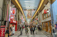 A bustling covered shopping street in Japan with people walking, colorful signs, hanging lanterns, and various shops, including a Sugakiya restaurant. A large floral banner hangs overhead.