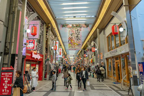 A bustling covered shopping street in Japan with people walking, colorful signs, hanging lanterns, and various shops, including a Sugakiya restaurant. A large floral banner hangs overhead.