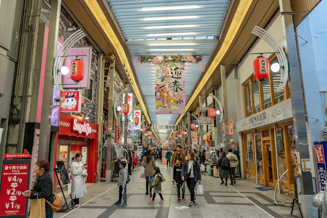 A bustling covered shopping street in Japan with people walking, colorful signs, hanging lanterns, and various shops, including a Sugakiya restaurant. A large floral banner hangs overhead.