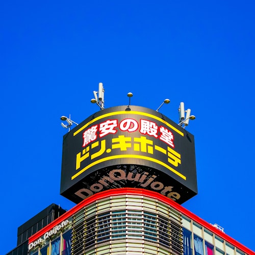 Don Quijote A Don Quijote store sign with yellow and red Japanese text is displayed atop a building against a clear blue sky. The top of the building features antennas and curved architectural elements.