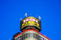 A Don Quijote store sign with yellow and red Japanese text is displayed atop a building against a clear blue sky. The top of the building features antennas and curved architectural elements.