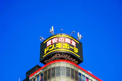 A Don Quijote store sign with yellow and red Japanese text is displayed atop a building against a clear blue sky. The top of the building features antennas and curved architectural elements.
