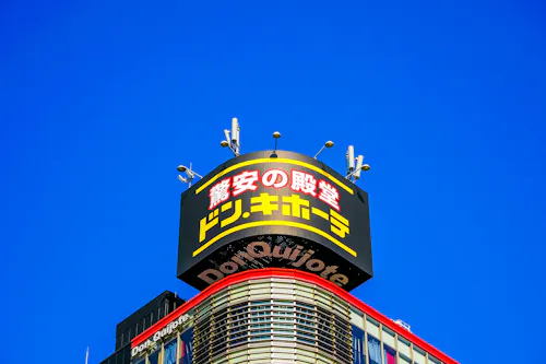 A Don Quijote store sign with yellow and red Japanese text is displayed atop a building against a clear blue sky. The top of the building features antennas and curved architectural elements.