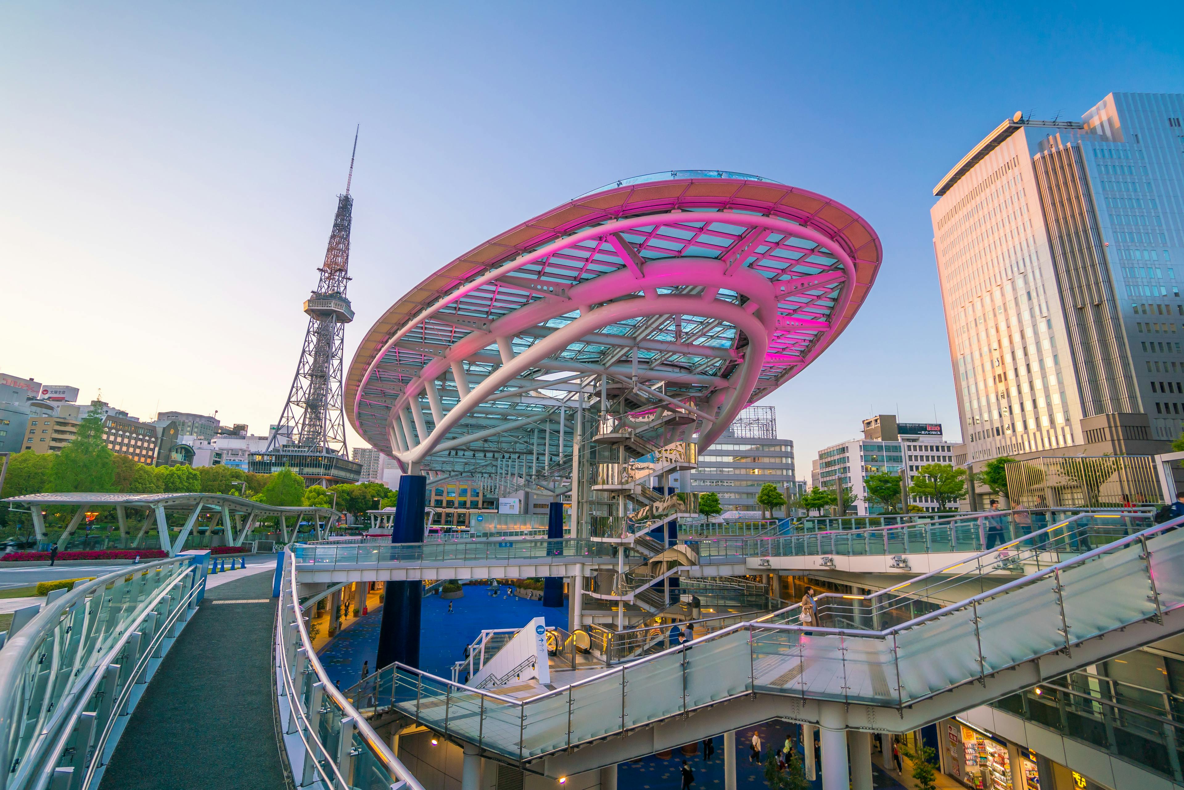 Futuristic glass dome structure with pink lighting at Oasis 21 in Nagoya, Japan, surrounded by modern buildings, walkways, and the Nagoya TV Tower in the background at sunset.