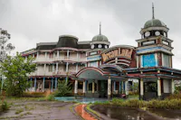 An abandoned, graffiti-covered building with towers and a sign reading “Dreamland,” surrounded by overgrown grass and cloudy skies, suggesting a deserted amusement park.
