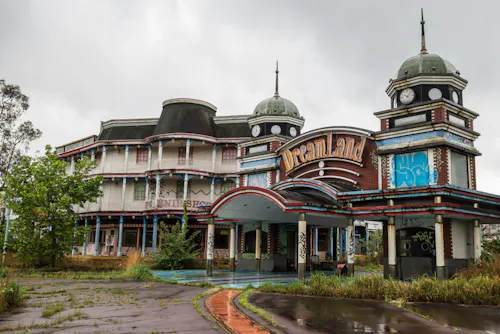 An abandoned, graffiti-covered building with towers and a sign reading “Dreamland,” surrounded by overgrown grass and cloudy skies, suggesting a deserted amusement park.