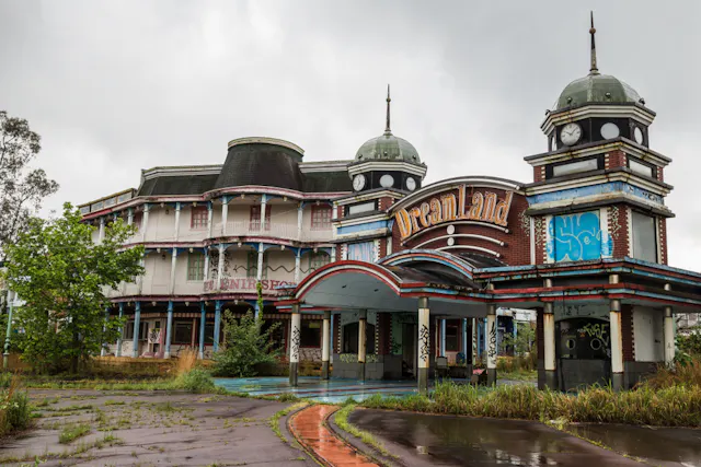 An abandoned, graffiti-covered building with towers and a sign reading “Dreamland,” surrounded by overgrown grass and cloudy skies, suggesting a deserted amusement park.