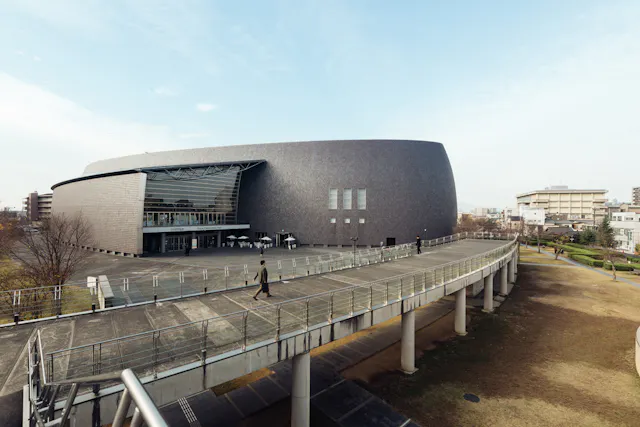 A modern, oval-shaped building with a dark exterior and large glass entrance sits beside a curved elevated walkway supported by columns, surrounded by grass and a few people walking nearby.