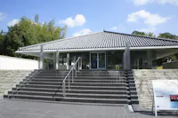 A modern building with a tiled, sloped roof and glass entrance stands behind wide stone steps and metal railings, surrounded by greenery and a blue sky with clouds. Signs are visible near the entrance.