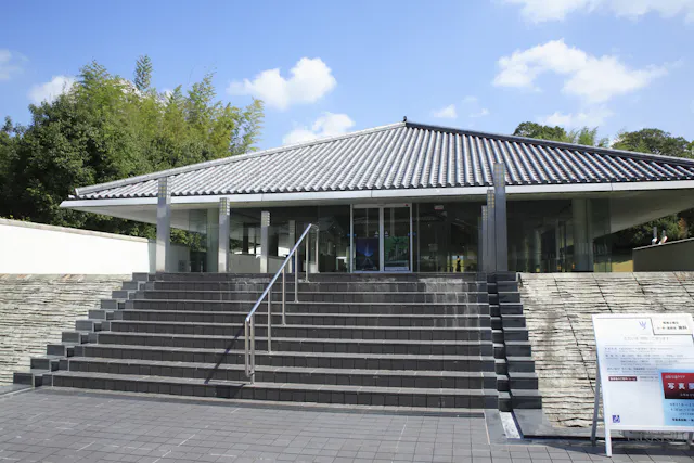 A modern building with a tiled, sloped roof and glass entrance stands behind wide stone steps and metal railings, surrounded by greenery and a blue sky with clouds. Signs are visible near the entrance.