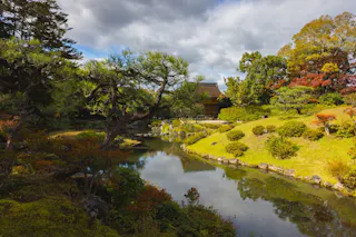 A tranquil Japanese garden with a pond, lush green grass, trees with autumn colors, and a traditional wooden building in the background under a partly cloudy sky.