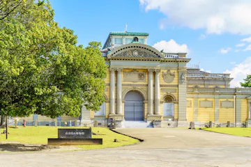 Nara National Museum A grand, historic yellow building with classical columns and arched windows stands in the sunlight, surrounded by green trees. A sign with Japanese writing is displayed near the entrance.