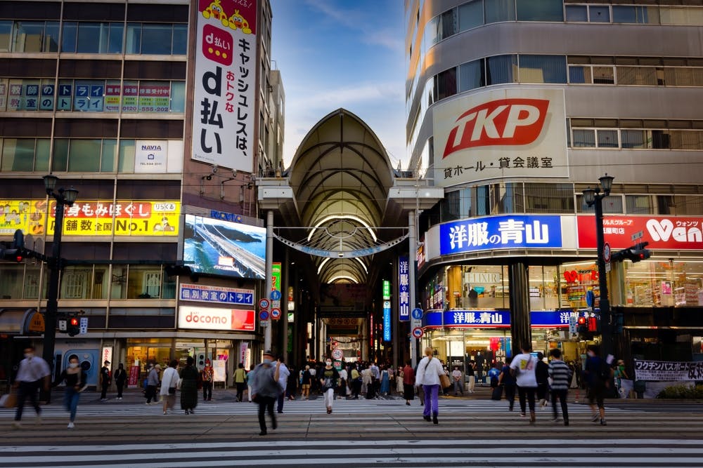Busy city street in Japan with people crossing a wide crosswalk, surrounded by tall buildings featuring bright signs and advertisements in Japanese. An arched shopping arcade entrance is visible in the background.