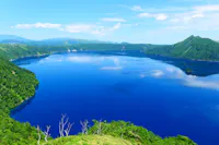 A deep blue lake surrounded by lush green hills under a clear blue sky with scattered clouds, reflecting in the calm water.