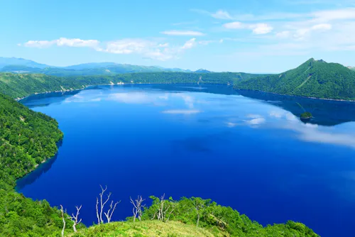 A deep blue lake surrounded by lush green hills under a clear blue sky with scattered clouds, reflecting in the calm water.