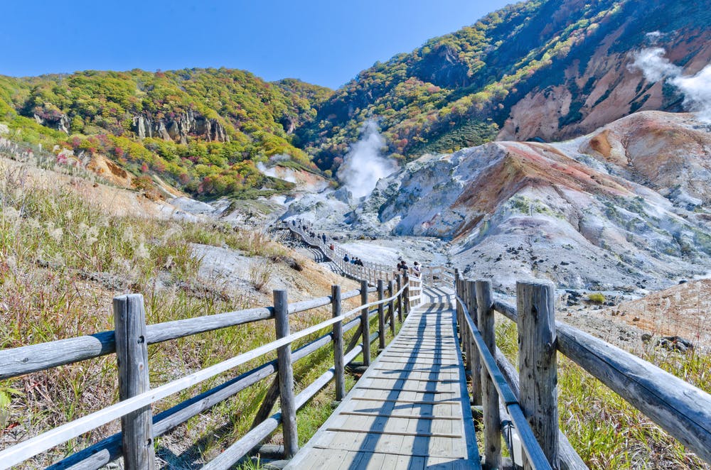 A wooden walkway leads through a rocky, steaming geothermal valley surrounded by green hills and colorful mineral deposits under a clear blue sky.