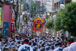 A large crowd participates in a Japanese festival parade, wearing matching traditional clothing and headbands, surrounding a decorated portable shrine as they move down a city street.