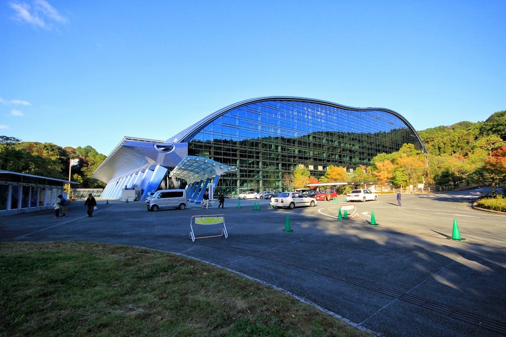 A large, modern building with a curved, glass facade reflects the blue sky and trees. Several cars are parked in front, with green traffic cones and a barrier on a paved area surrounded by greenery.